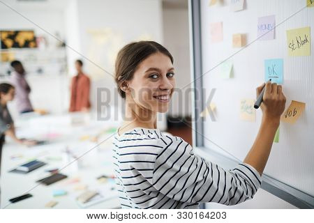 Waist Up Portrait Of Youngwoman Looking At Camerawhile Writing On Whiteboard In Office, Copy Space
