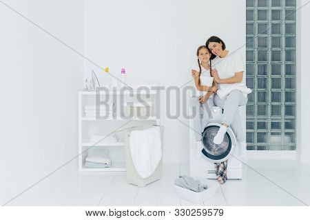 Shot Of Beautiful Woman And His Small Daughter Embrace And Smile Pleasantly, Sit On Washing Machine,