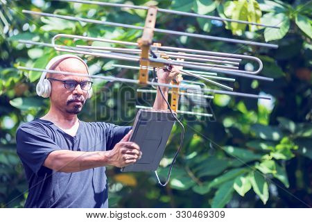 An Asian Man Is Carrying An Antenna And A Tablet In His Hands With Headphone To Find The Signal On G