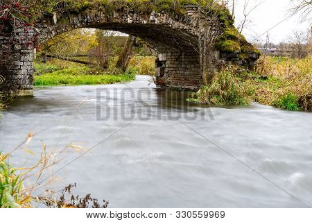 Old Stone Bridge Over A Small Fast River. Time Is Mercilessly In Man S Buildings And Slowly Destroys