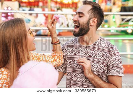 Image of a smiling positive loving couple walking outdoors in amusement park eat candyfloss.