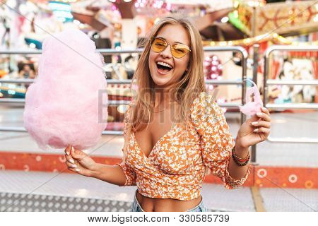 Photo of a positive cute woman walking outdoors in amusement park eat candyfloss.