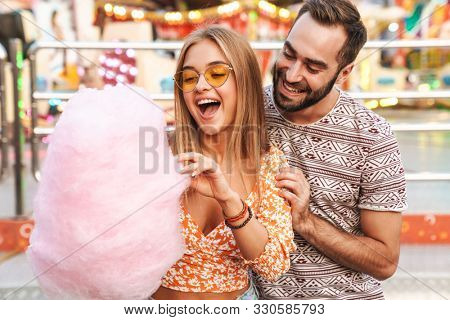 Image of a smiling positive loving couple walking outdoors in amusement park eat candyfloss.