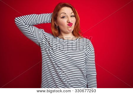 Young redhead woman wearing strapes navy shirt standing over red isolated background confuse and wonder about question. Uncertain with doubt, thinking with hand on head. Pensive concept.