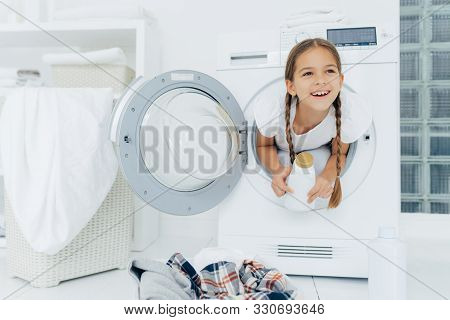 Happy European Girl With Two Plaits, Poses Inside Of Wash Machine, Holds White Bottle With Liquid Po