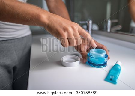 Man applying cream morning skincare regimen moisturizing face skin at bathroom mirror. Male beauty men skin care closeup of product jar and finger putting lotion.