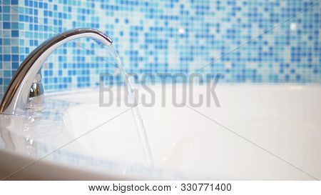 Close-up. Water Flows From A Luxurious Water Tap Into A Large Modern Bathtub Against A Blue Mosaic W
