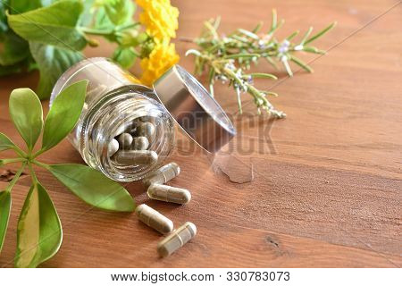 Natural Medicine Capsules In Open Glass Jar Lying On Wood Table With Flowers And Plant Leaves. Natur