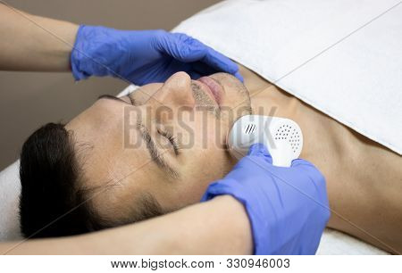Young Man Laying On A Facial Therapy Couch Covered With The White Towel With Opened Eyes. Facial The