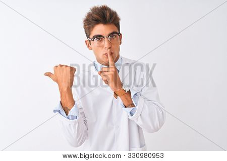 Young handsome sciencist man wearing glasses and coat over isolated white background asking to be quiet with finger on lips pointing with hand to the side. Silence and secret concept.