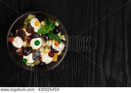 Salad from grapes apples pears kiwi oranges with mascarpone chease and cream. Healthy fresh fruit summer salad in glass bowl on black wooden background with copy space.