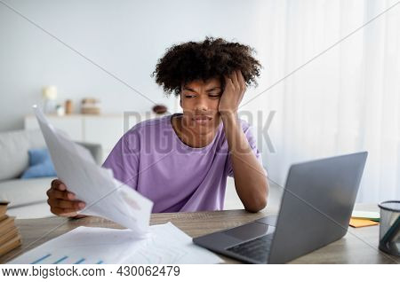 Bored Teen Sitting At Desk With Documents, Using Laptop To Write Coursework Paper, Studying Online, 