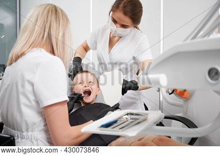 Adorable Little Boy Sitting In Dental Chair While Two Female Dentists Checking Kid Teeth. Dentist Ex