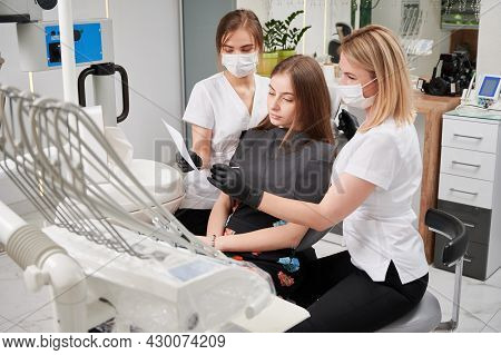 Beautiful Young Woman Sitting In Dental Chair While Stomatologists In Medical Masks Analyzing Dental