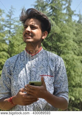Closeup Portrait Of A Indian Good Looking Young Guy Standing Outdoor In Nature And Holding Mobile Ph