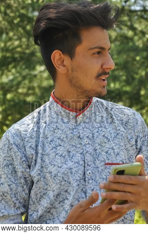 Closeup Portrait Of A Indian Good Looking Young Guy Standing Outdoor In Nature And Holding Mobile Ph