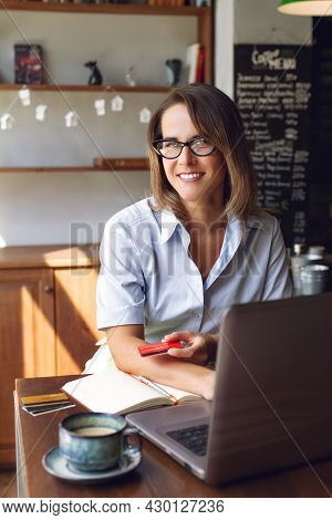 Portrait Of Smiling Woman In Glasses And Blue Shirt Sitting At Table With Laptop And Coffee Holding 