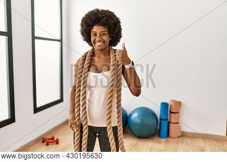 Young african american woman with afro hair at the gym training with battle ropes doing happy thumbs up gesture with hand. approving expression looking at the camera showing success. 
