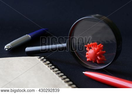 Red Toy Virus Under A Magnifying Glass On A Black Background Next To A Notebook And Pen. The Concept