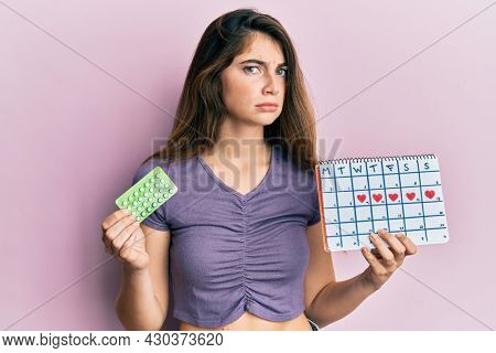 Young caucasian woman holding birth control pills and menstrual calendar skeptic and nervous, frowning upset because of problem. negative person. 
