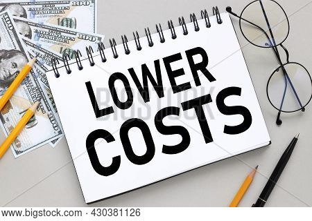 Lower Costs. Notepad On A Gray Background Near Bills And Pencils
