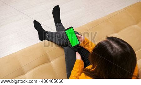 Long Haired Brunette In Jeans And Pullover Sits On Sofa Of Brown Colour And Types On Smartphone With