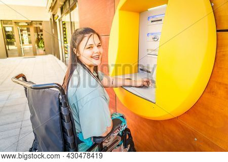 Young Happy Disabled Handicapped Woman In Wheelchair Using Atm Cash Macjine And Smiling At Camera On