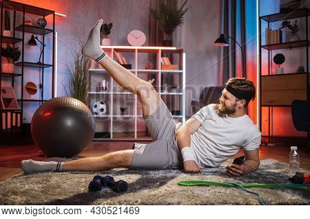 Young Positive In Good Mood Bearded Sportsman Lying On The Floor And Doing Leg Lifting Exercises Wit