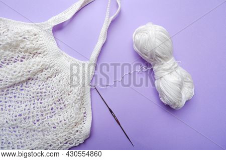 Eco Bag In The Knitting Process. White Grid Bag, Crochet Hook And Remaining Yarn On A Purple Backgro