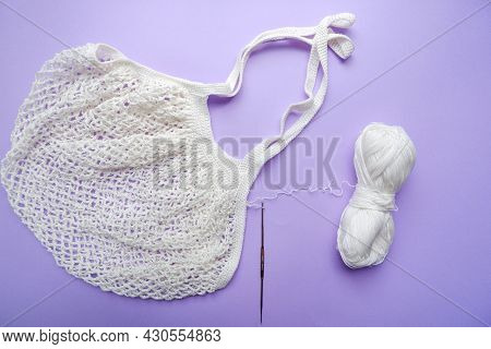 Eco Bag In The Knitting Process. White Grid Bag, Crochet Hook And Remaining Yarn On A Purple Backgro