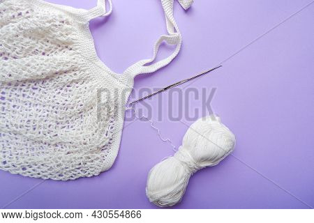 Eco Bag In The Knitting Process. White Grid Bag, Crochet Hook And Remaining Yarn On A Purple Backgro