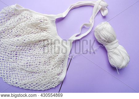 Eco Bag In The Knitting Process. White Grid Bag, Crochet Hook And Remaining Yarn On A Purple Backgro