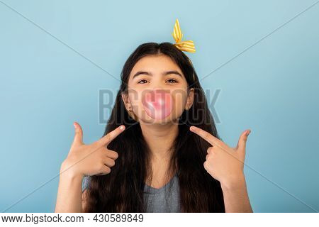 Funny Indian Teen Girl Blowing Bubble From Sugar Free Chewing Gum, Pointing At It Over Blue Studio B
