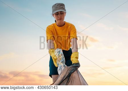 Portrait Of Female Volunteer In Rubber Gloves Puts A Dirty Plastic Bottle In Bag With Garbage. Sunse