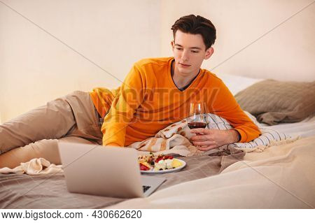 Relaxed Young Guy With Glass Of Wine In His Hands Laying On Comfortable Bed With Plate Of Food, Watc