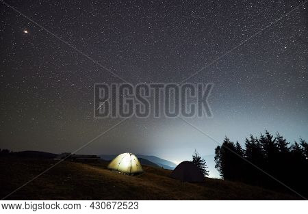 Night Camping In The Mountains. Two Tents, One Of Which Illuminated, Set Up On Mountain Meadow. Pano