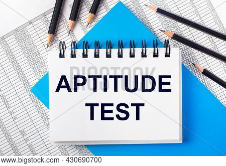 White Notebook With The Text Aptitude Test On The Table Next To Black Pencils On A Blue Background A