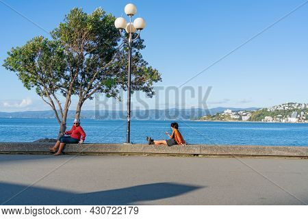 Wellington New Zealand - July 30 2021; Two Urban Women Using Device While Relaxing At Waters Edge On