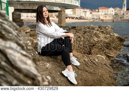 Young And Attractive Brunette In White Shirt Is Sitting On Rocky Seacoast. Woman Was Tired Of Her Fo