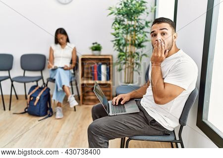 Young people sitting at waiting room working with laptop covering mouth with hand, shocked and afraid for mistake. surprised expression 