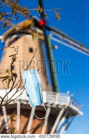 Covert-19 Face Mask Left Hanging On A Tree Branch. With Foxton Windmill In The Background.