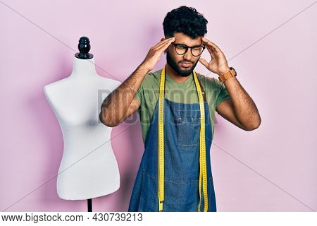Arab man with beard dressmaker designer wearing atelier apron with hand on head, headache because stress. suffering migraine. 