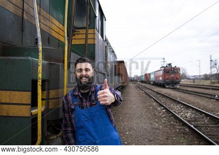 Portrait Of Engine Train Driver Standing By Locomotive At Train Station And Holding Thumbs Up.