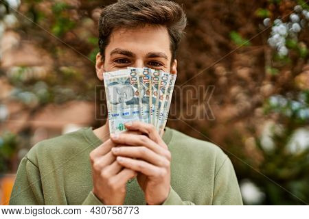 Young hispanic man smiling happy covering face with peruvian soles banknotes at the city.