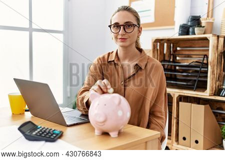 Young blonde woman at the office putting coin into piggy bank thinking attitude and sober expression looking self confident 