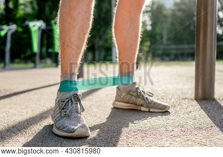 Closeup front view of legs feets of man guy training with elastic rubber band at the stadium outdoors. Athlete adult male person during active workout with additional sport equipmant in summertime
