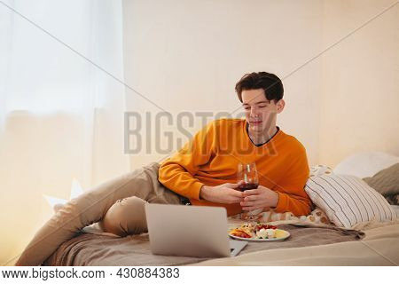 Relaxed Young Guy With Glass Of Wine In His Hands Laying On Comfortable Bed With Plate Of Food, Watc