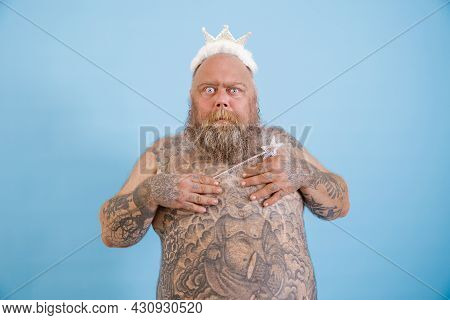 Funny Obese Man With Toy Crown And Magic Stick Covers Breasts On Light Blue Background