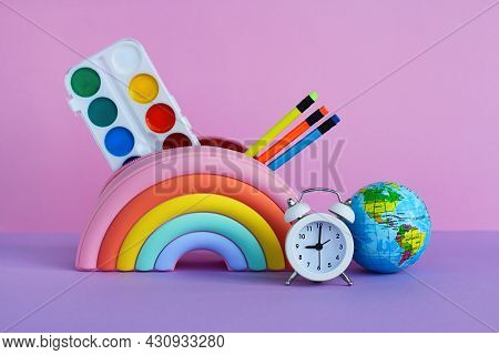 A Pencil Case In The Form Of A Bright Rainbow With Paints, Pencils, A White Alarm Clock And A Toy Pl