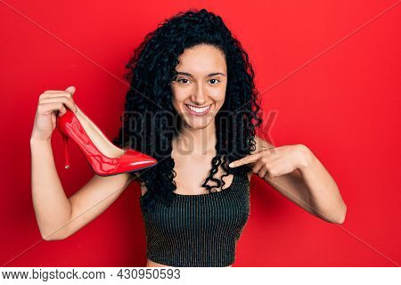 Young hispanic woman with curly hair holding red high heel shoes pointing finger to one self smiling happy and proud 